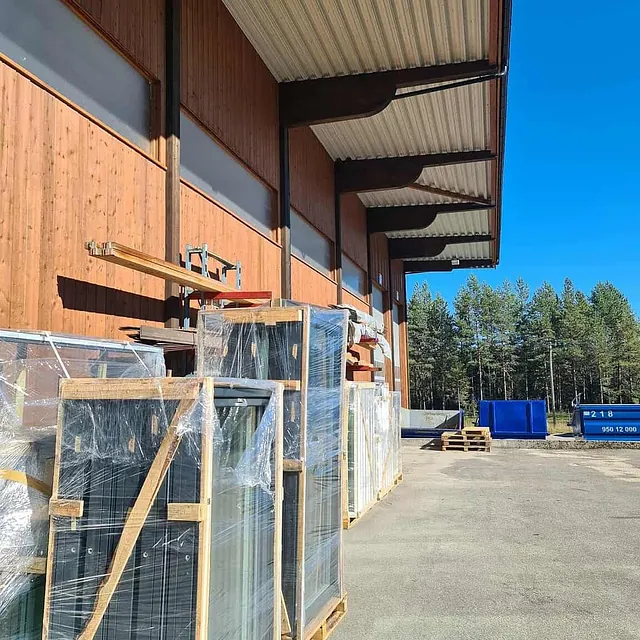 Industribygg med trekledning og materialer lagret utendørs langs vegg i sollys.